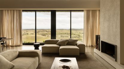 A bright, airy beige living room with sectional sofas, a wood side table, a fireplace, a nook for firewood, and sheer curtains