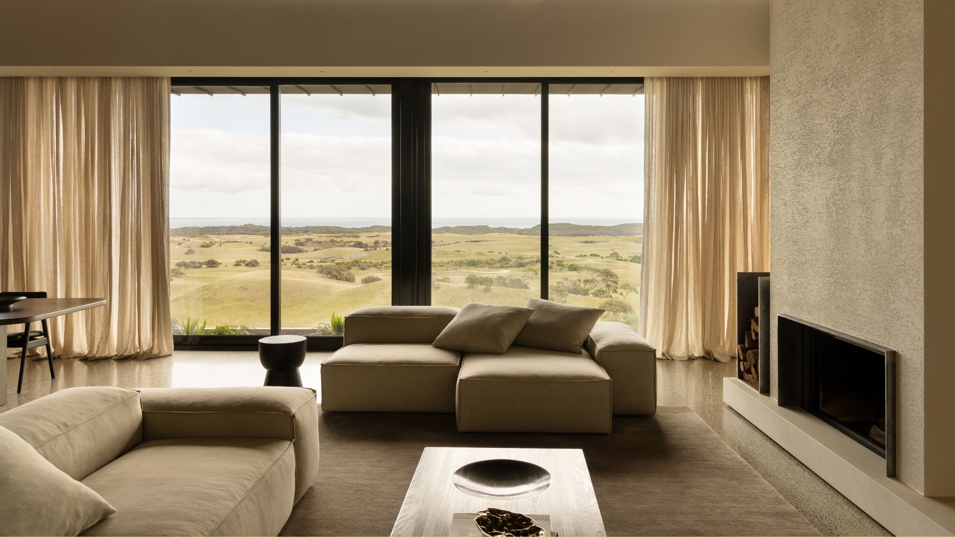 A bright, airy beige living room with sectional sofas, a wood side table, a fireplace, a nook for firewood, and sheer curtains