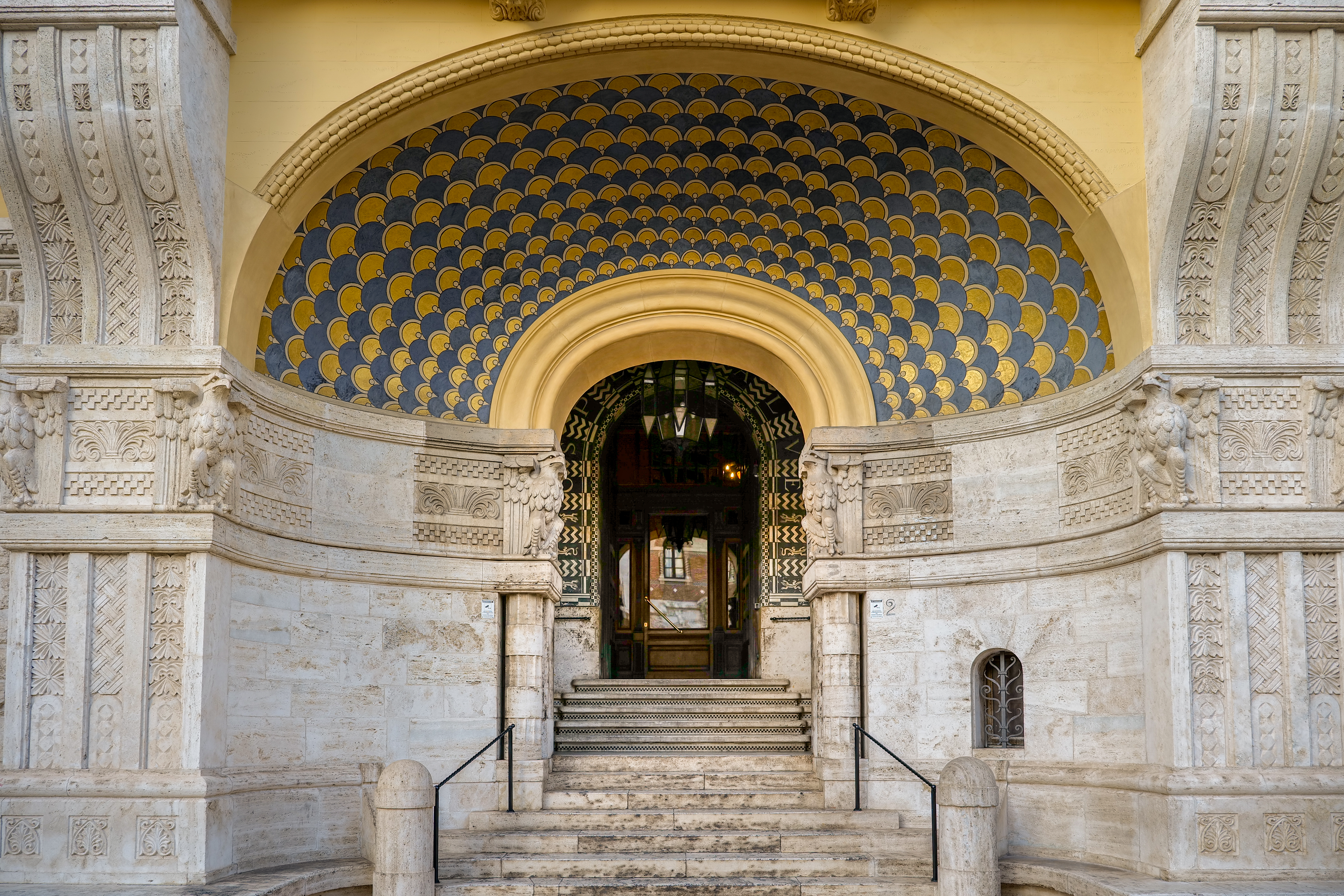 The entrance to an intricately designed building with limestone, sculpted exterior and an arch decorated with an eye-catching vault in blue and golden leaf-shaped decorations.