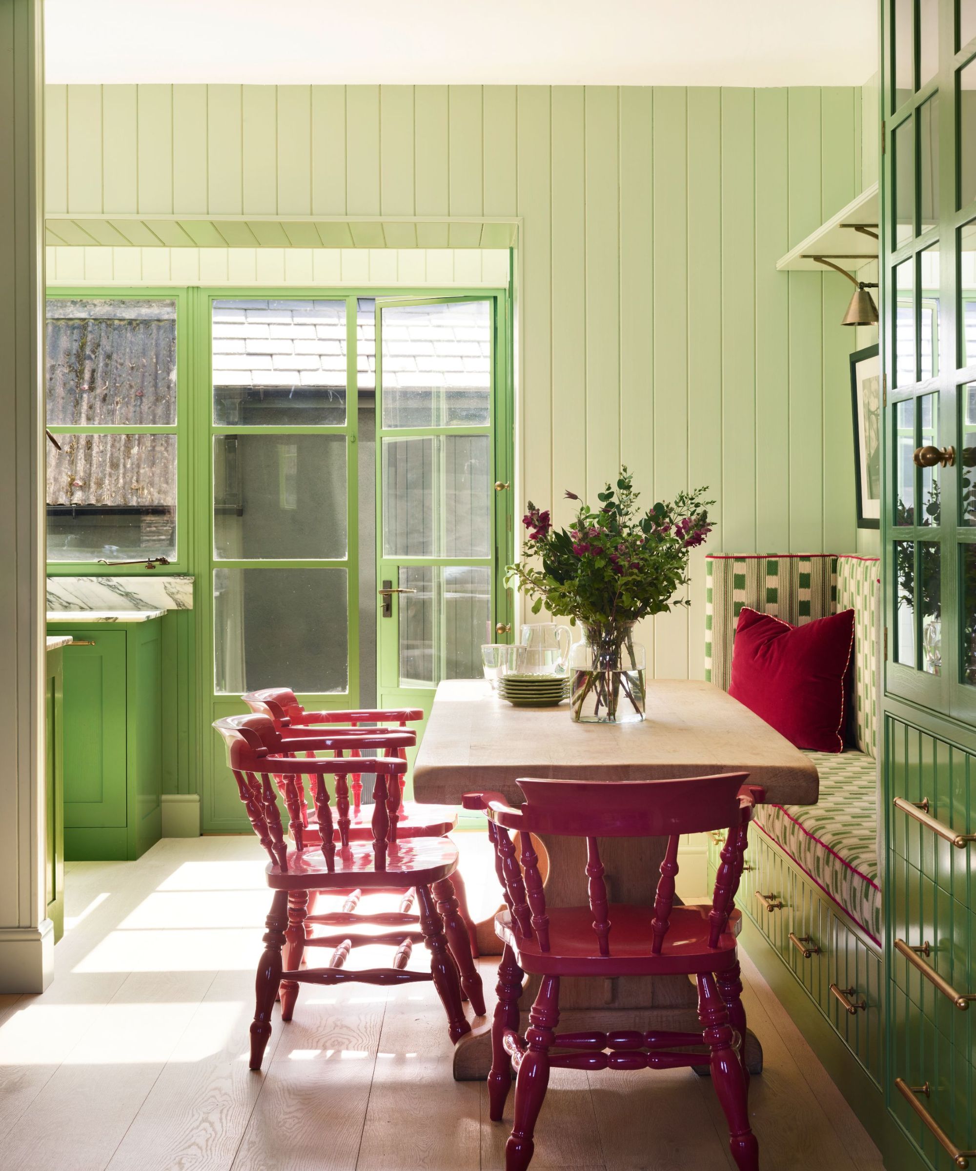 A green kitchen with a dining nook featuring a rectangular wooden table, three antique red dining chairs, and an upholstered banquette