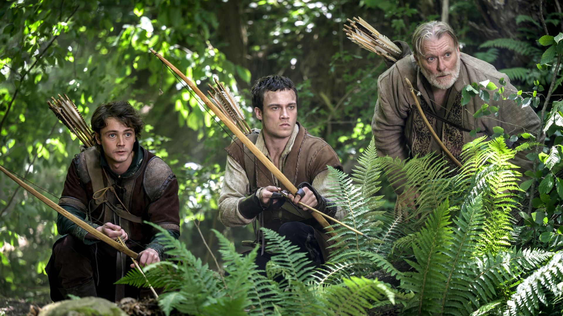Robin Hood and two Merry Men draw their bows while hiding in the woods