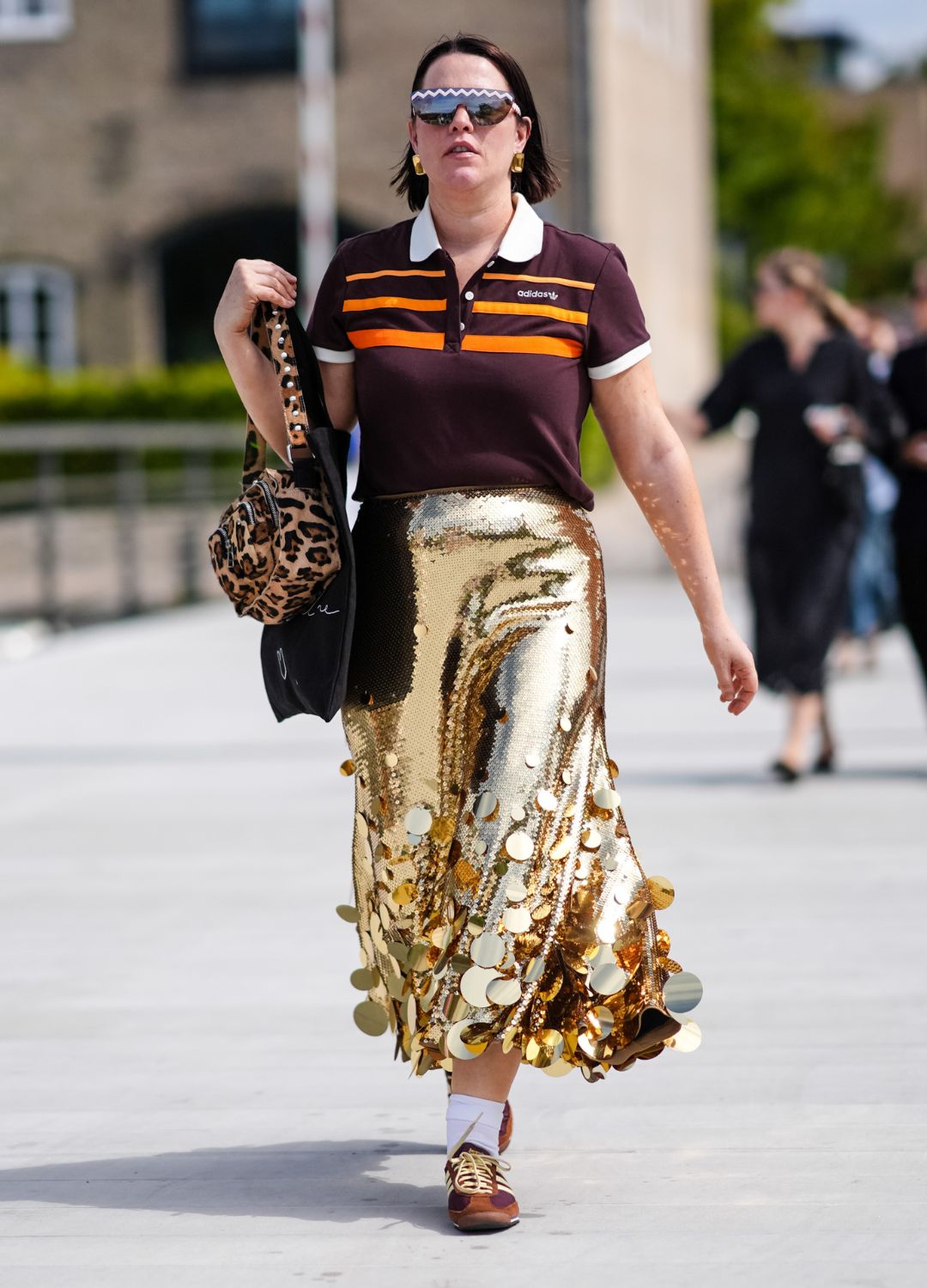 A guest wears black sunglasses, gold earrings, dark red brown bright orange striped Adidas Originals 80S short sleeve shirt, light brown and dark brown leopard pattern/print bag, shiny gold sequin with circle detail midi skirt, white socks, dark brown Adidas Originals sambas SL-72, outside Marimekko, during the Copenhagen Fashion Week Spring/Summer 2024-2025 on August 8, 2024 in Copenhagen, Denmark
