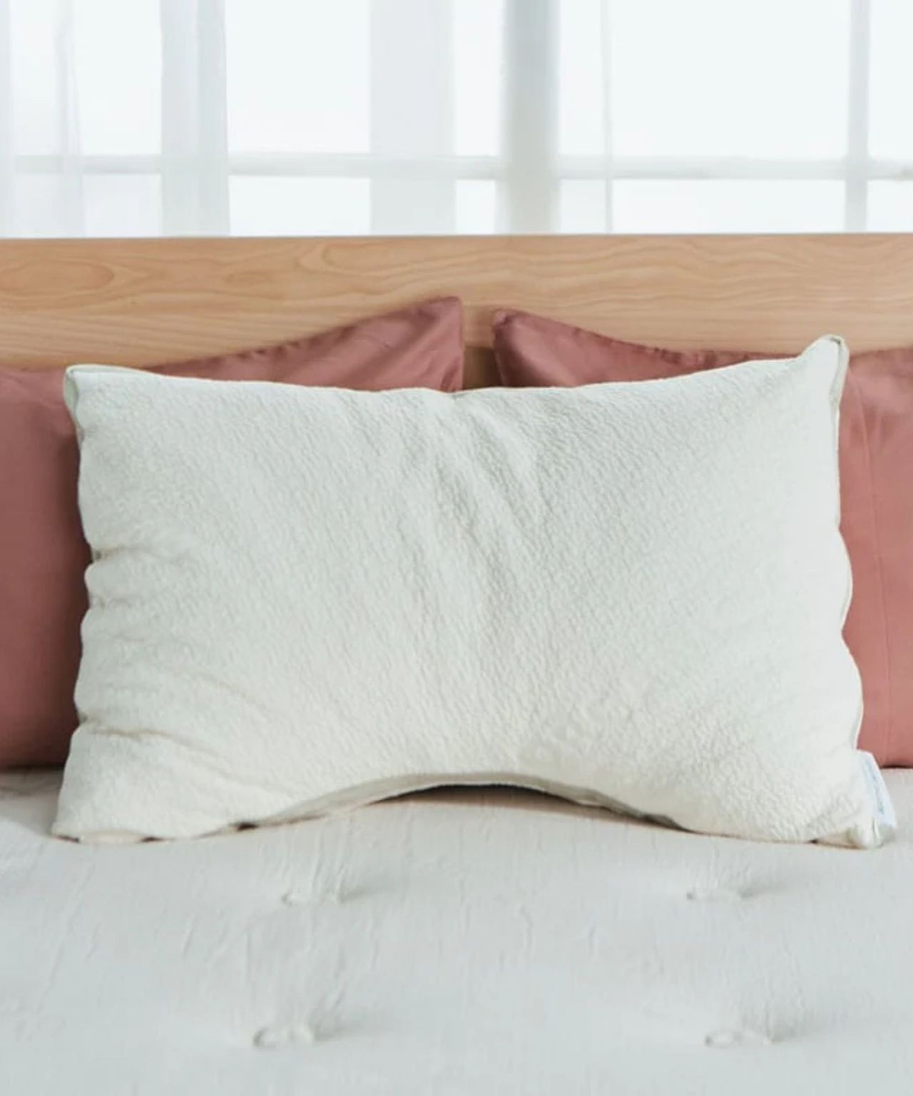 A white Avocado side sleeper green pillow pictured on a white mattress with salmon colored pillows behind and a light wood headboard with windows behind.