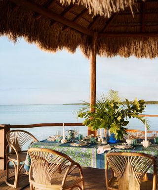 An outdoor dining table on the beach with a large floral arrangement and a green and blue tablecloth