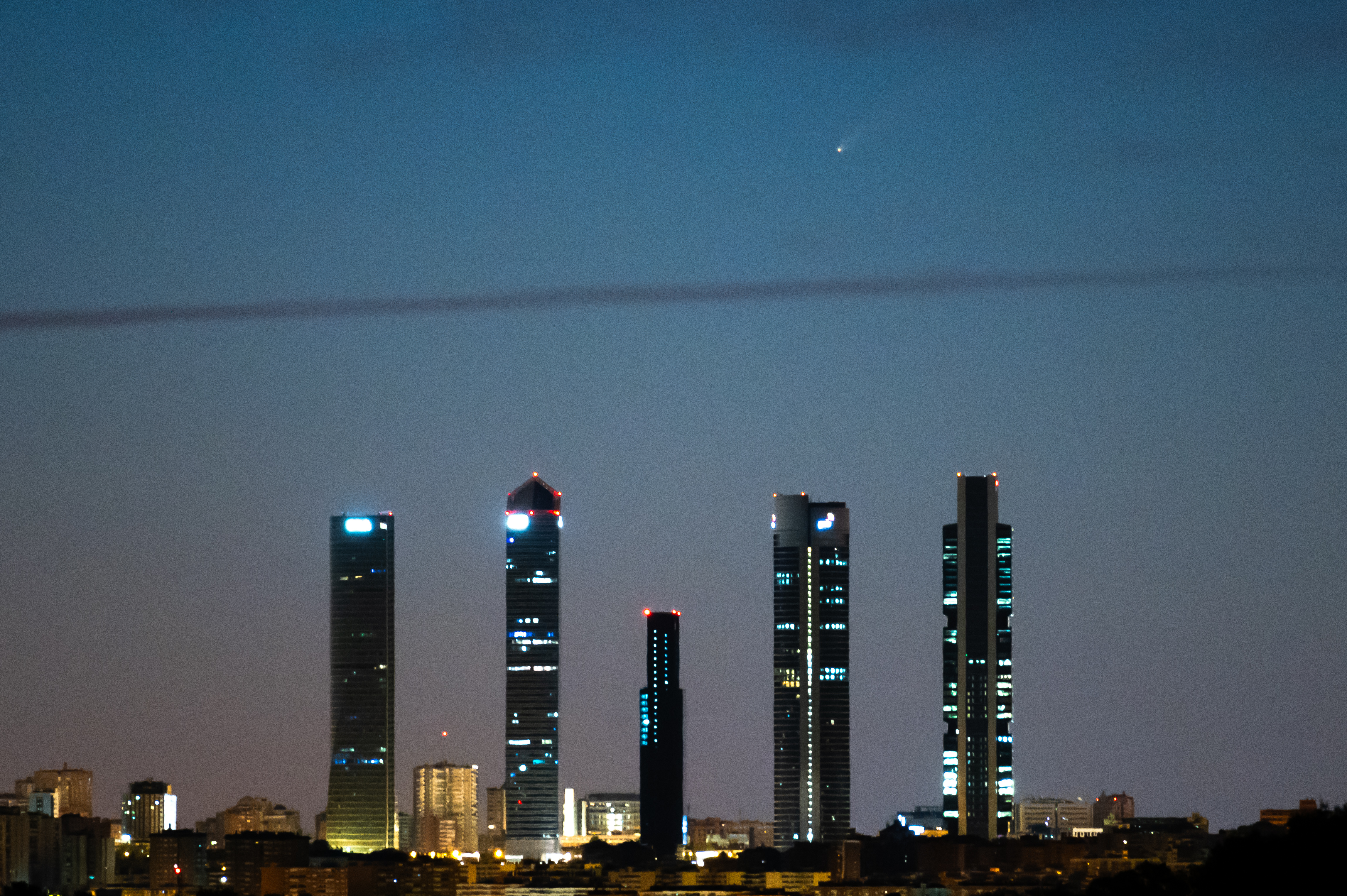 the faint streak of the comet tail is visible in the early morning sky above five large skywscrapers.