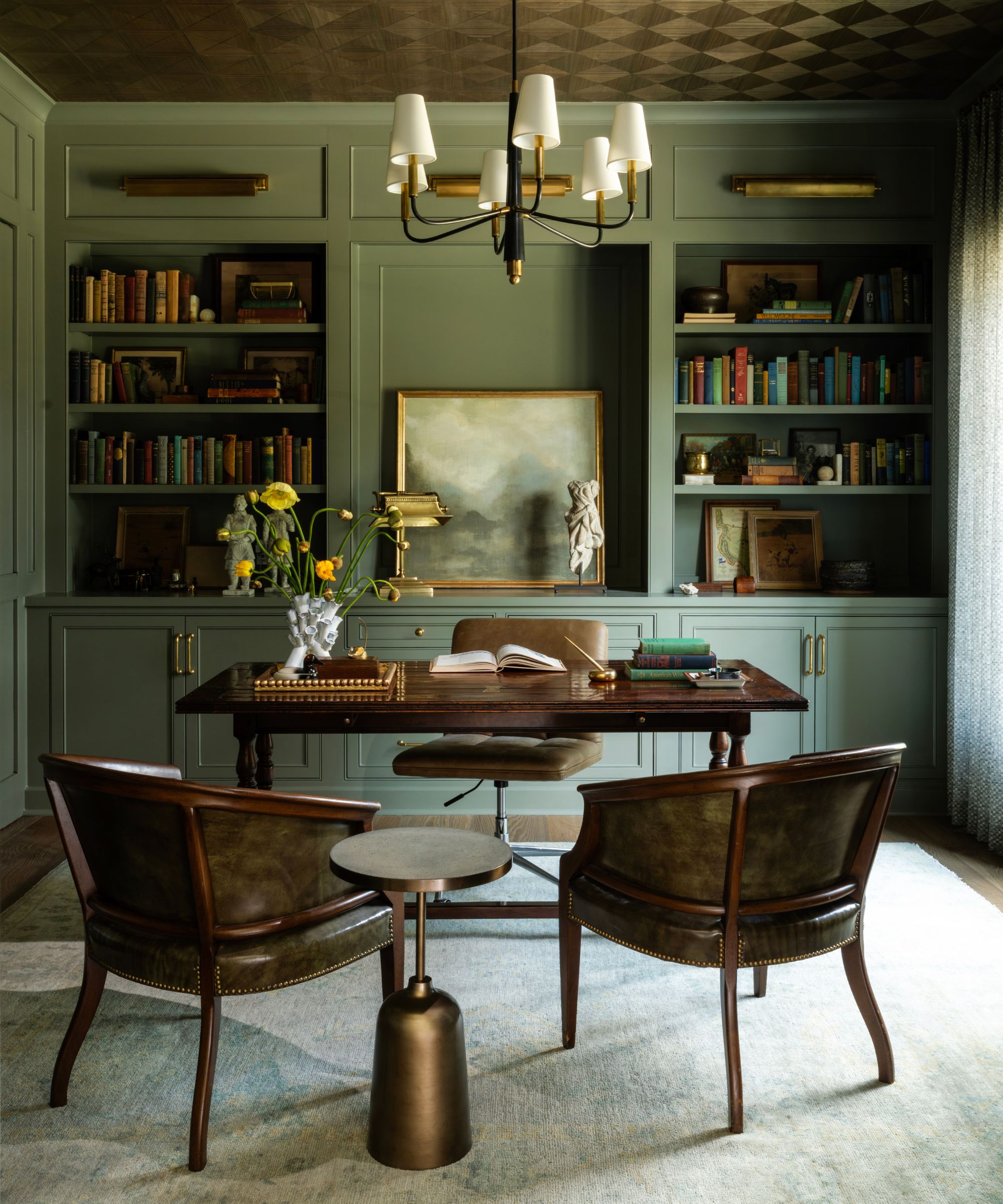 Home office with green painted bookshelves and cabinetry, a tiled ceiling, a candelabra light, a wooden desk and a leather desk chair, and two curved back armchairs.