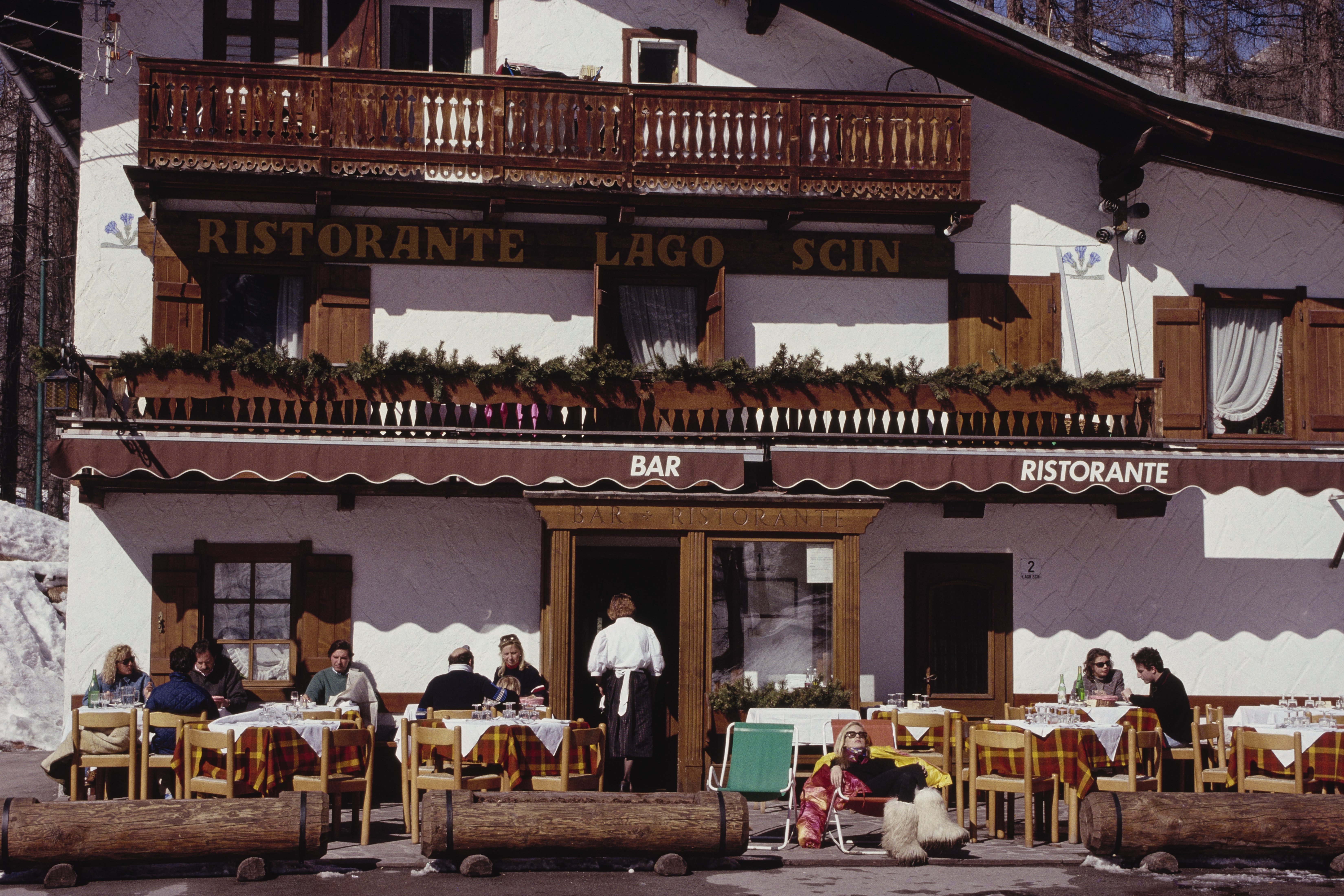 A traditional alpine restaurant with wooden balconies and outdoor tables, where diners sit in the winter sun surrounded by snow.