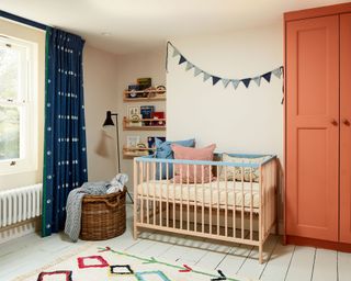 nursery with orange painted wardrobe and blue accents, light wood crib with bunting and cushions. weaved laundry basket and rug on painted white wood floor