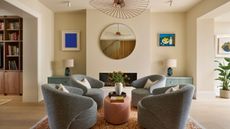 A modern living room with cream walls, a set of four gray armchairs in the middle of the room with a terracotta upholstered coffee table, an orange flecked rug and a modern pendant light. 