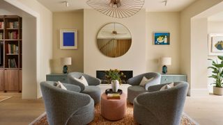 A modern living room with cream walls, a set of four gray armchairs in the middle of the room with a terracotta upholstered coffee table, an orange flecked rug and a modern pendant light. 