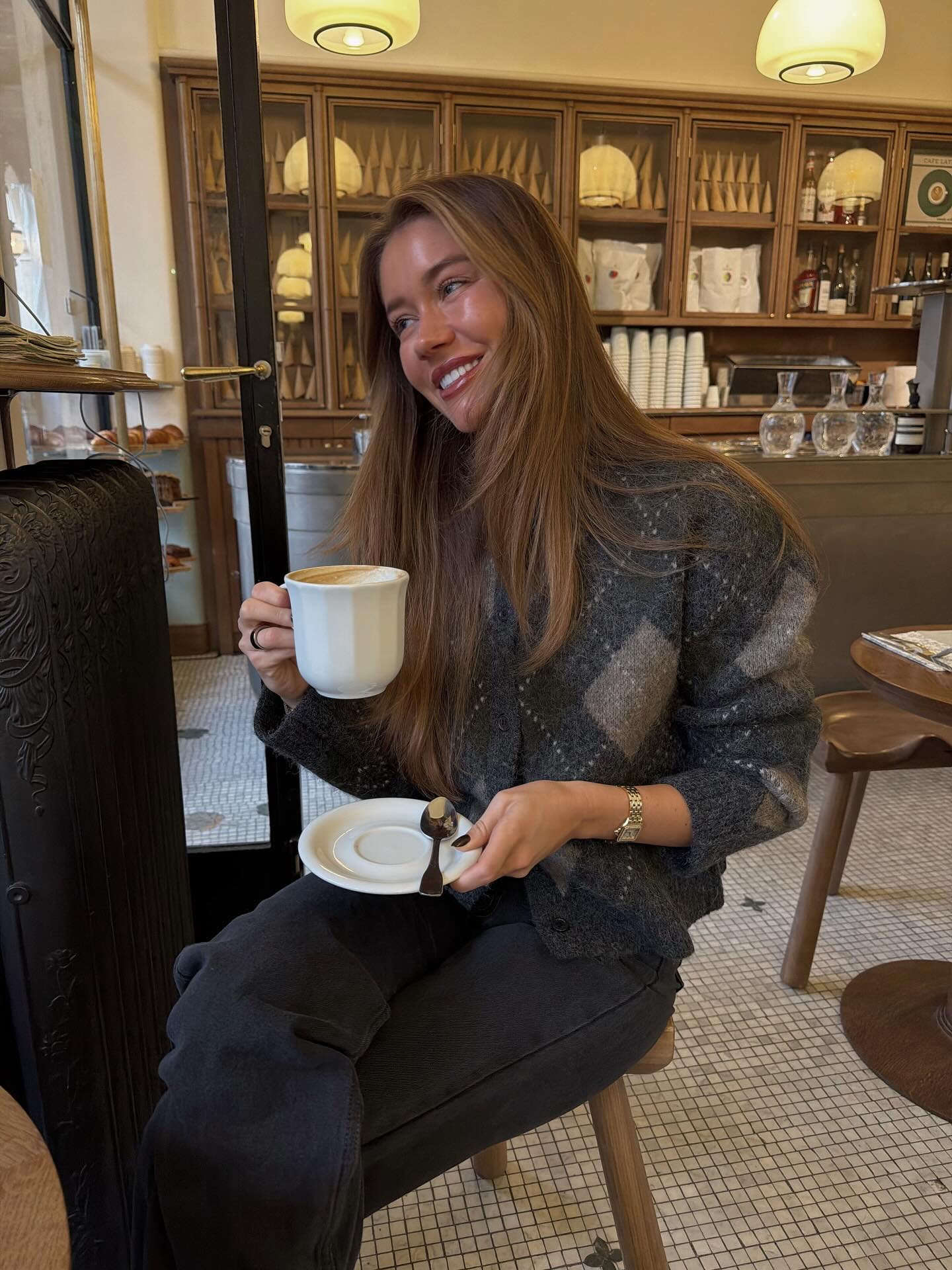 An influencer in Paris wearing an argyle cardigan and black jeans in a cafe wearing a silver watch