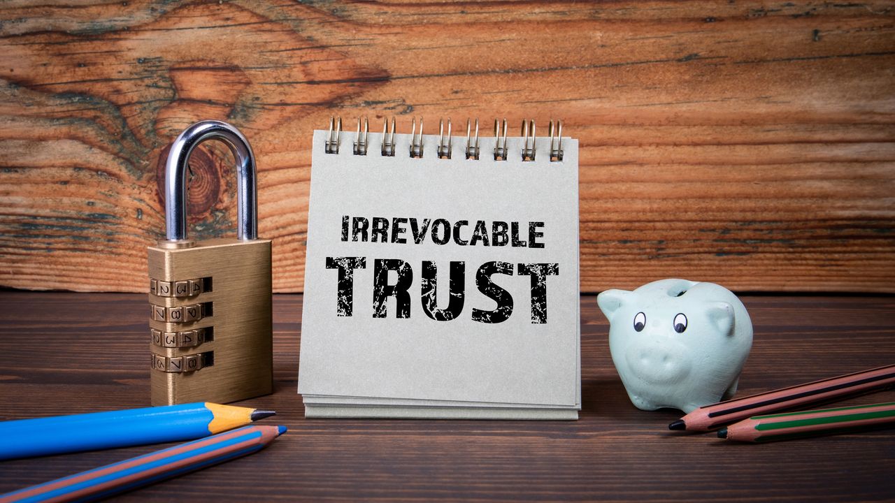 A spiral notebook displays the words &quot;irrevocable trust&quot; flanked by a padlock and a small piggy bank.