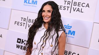 Image of Demi Moore smiling with long curly black hair and wearing a white sleeveless shirt