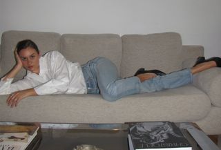 Anne-Laure Mais wearing a white button-down shirt, Levi's jeans, and ballet pumps while laying on a sofa inside her apartment.