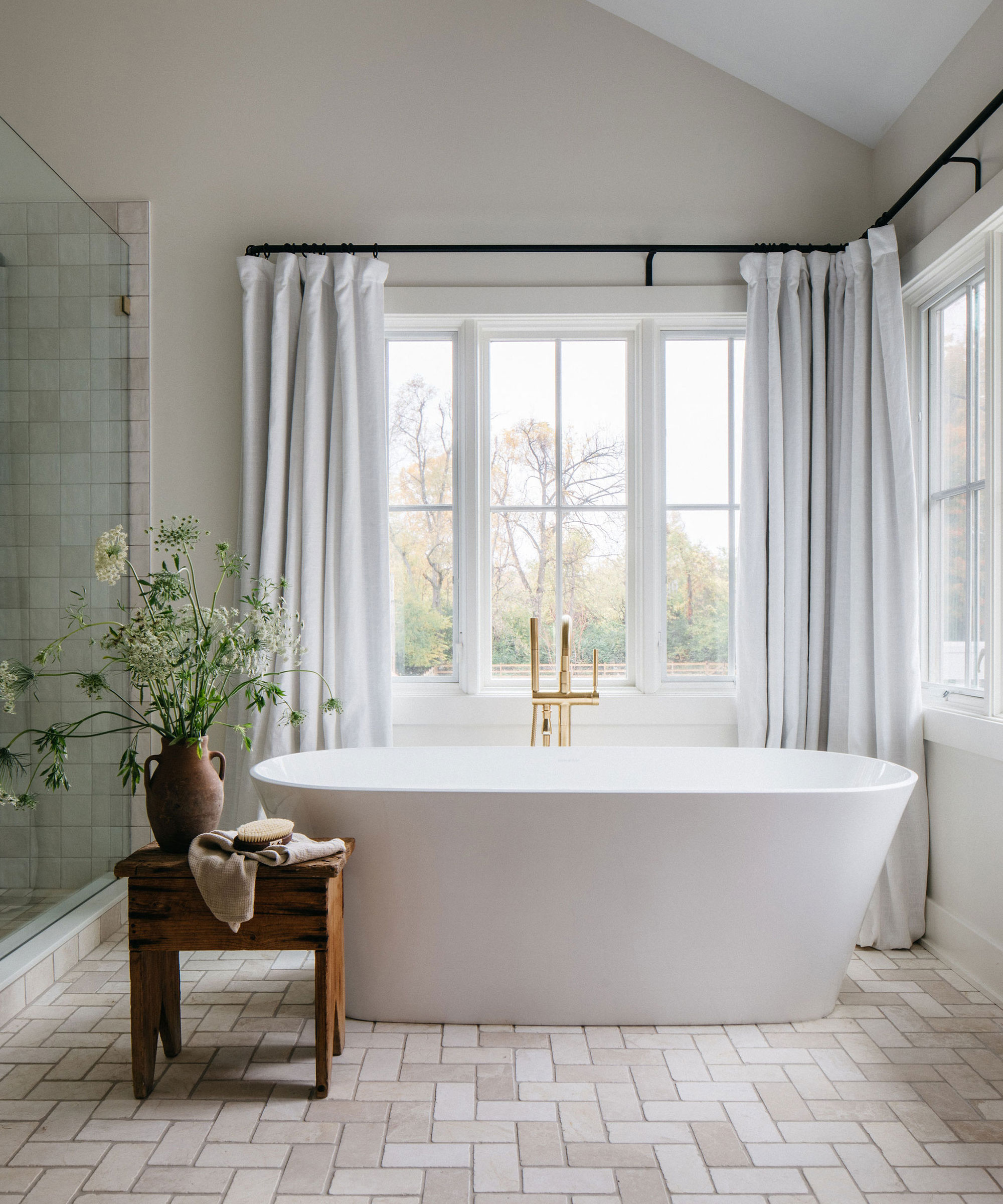 Neutral bathroom with white tub, matching square floor and shower tile and light drapes with landscape setting beyond window