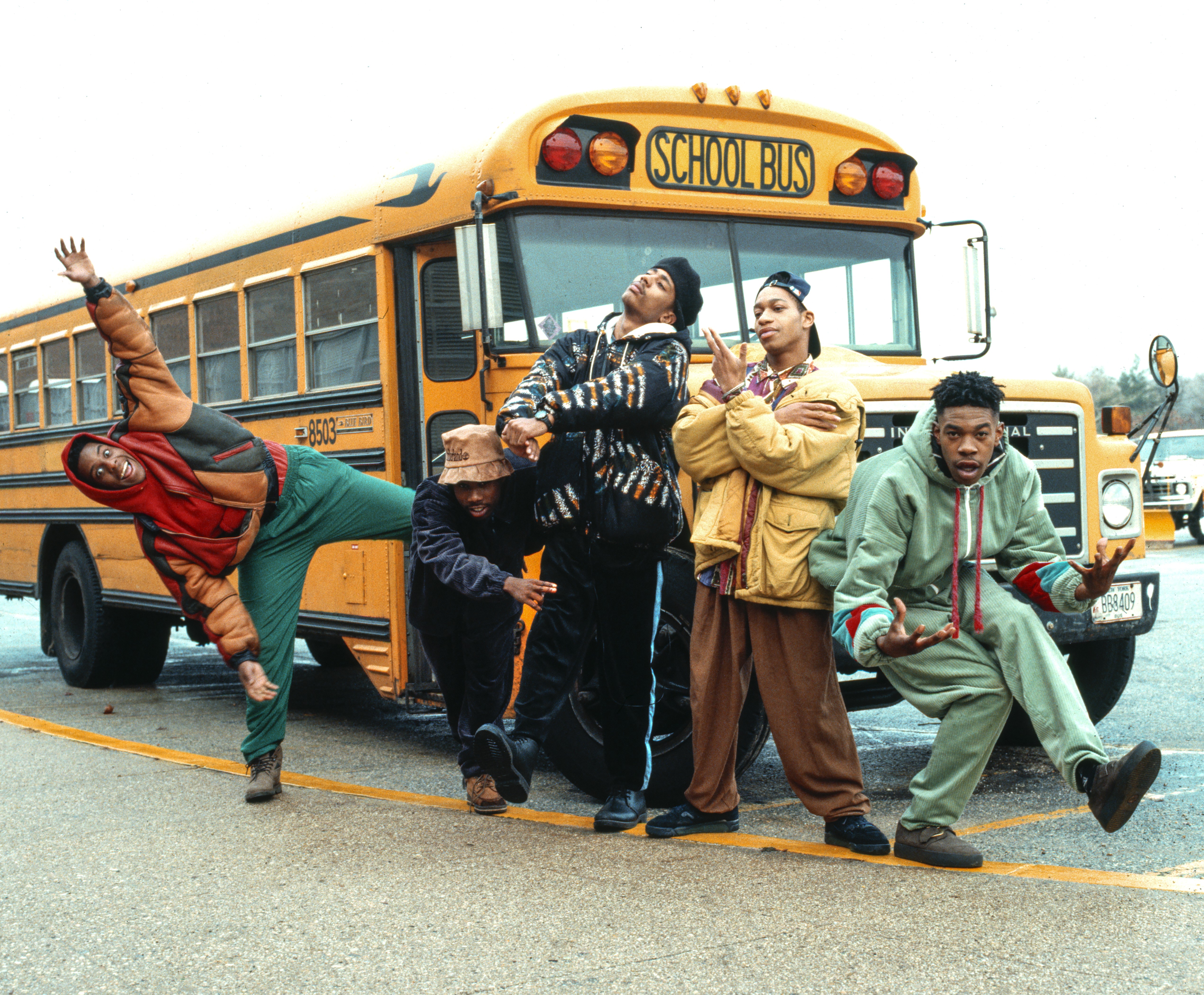 Five young men in colorful 1990s-style streetwear strike various energetic poses in front of a classic yellow school bus.
