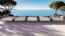 Five white Saatva mattresses lined up in front of a seascape with trees, bright blue sea, and blue skies.