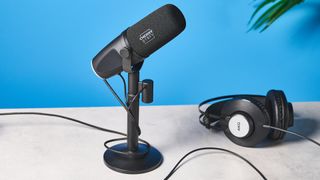 a black microphone by cherry XTRFY with a branded muff and an XLR and USB-C port sits atop a pale brown desk with a dark blue background