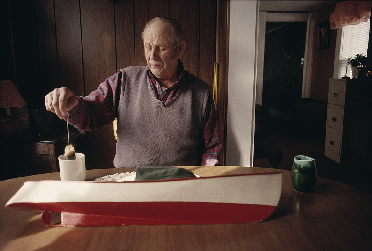 an elderly individual sat at a table removing a teabag from a mug