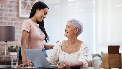 A standing young woman smiles down at an older woman sitting in front of an open laptop.