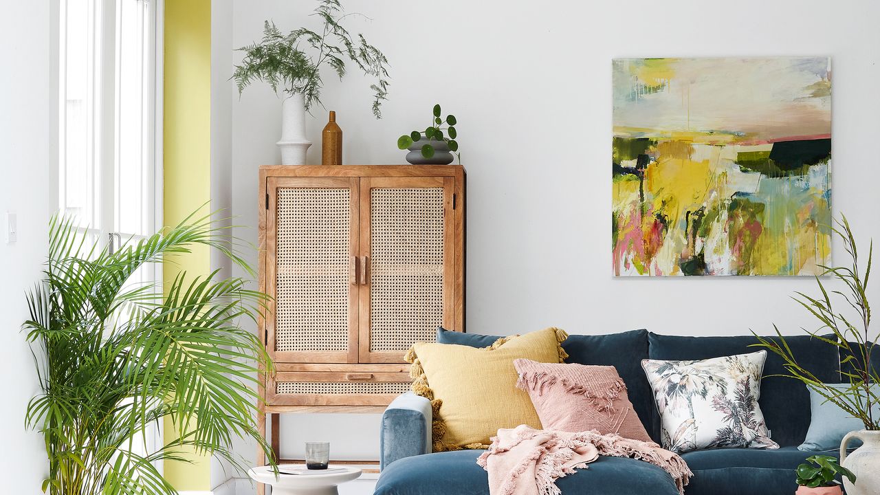 White living room with blue l-shaped sofa, rattan fronted cabinet and yellow painted architrave