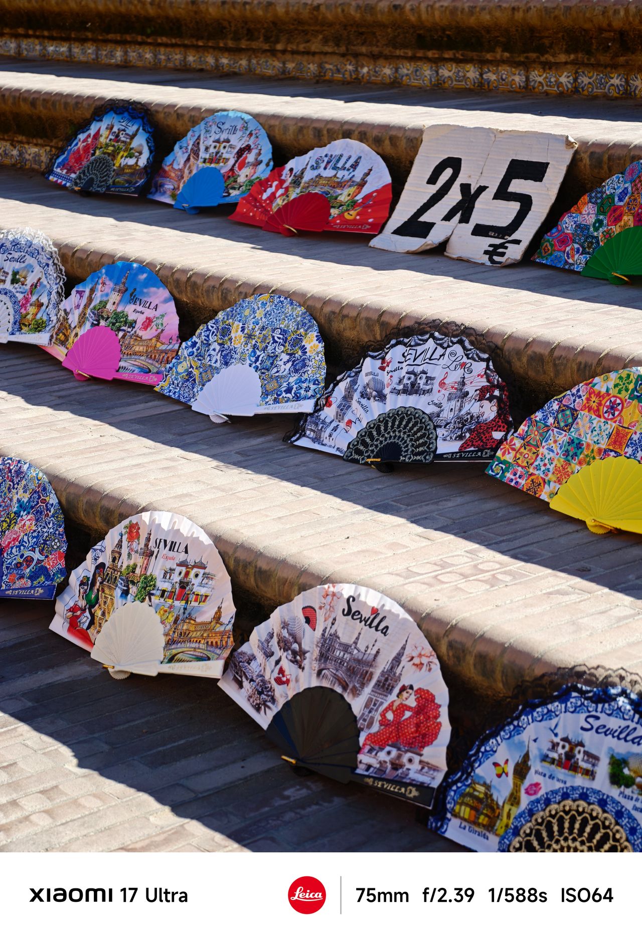 Colorful souvenir hand fans displayed on stone steps with a handwritten sign reading &ldquo;2x5&euro;&rdquo; in bright sunlight.