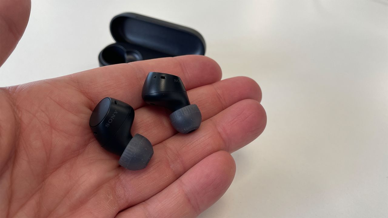 A hand holding a pair of black Sony WF-C510 in-ear headphones. The open charging case can be seen in the background.