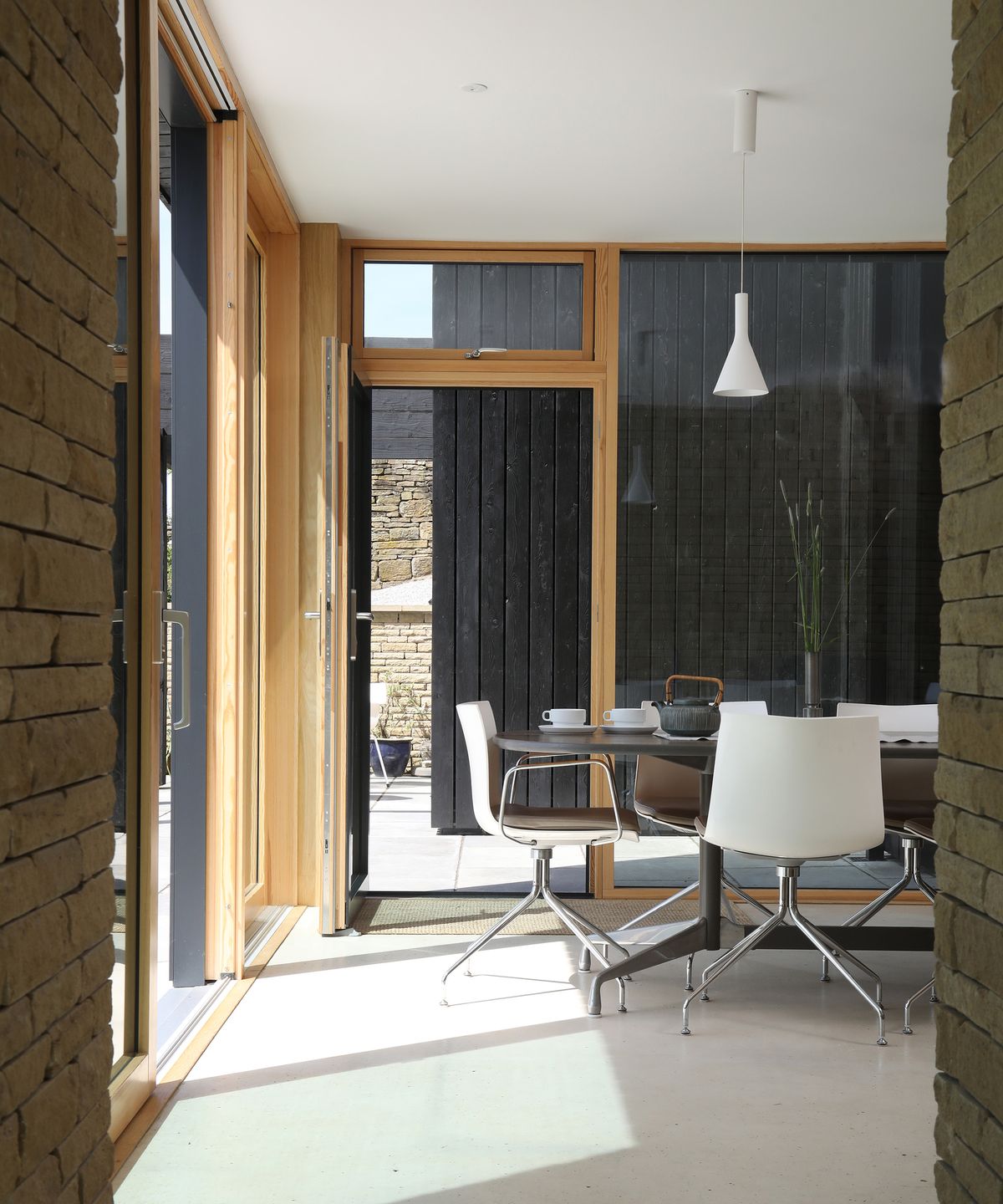 A dining table in a self build home with large glazing which looks through to a second building clad in black timber