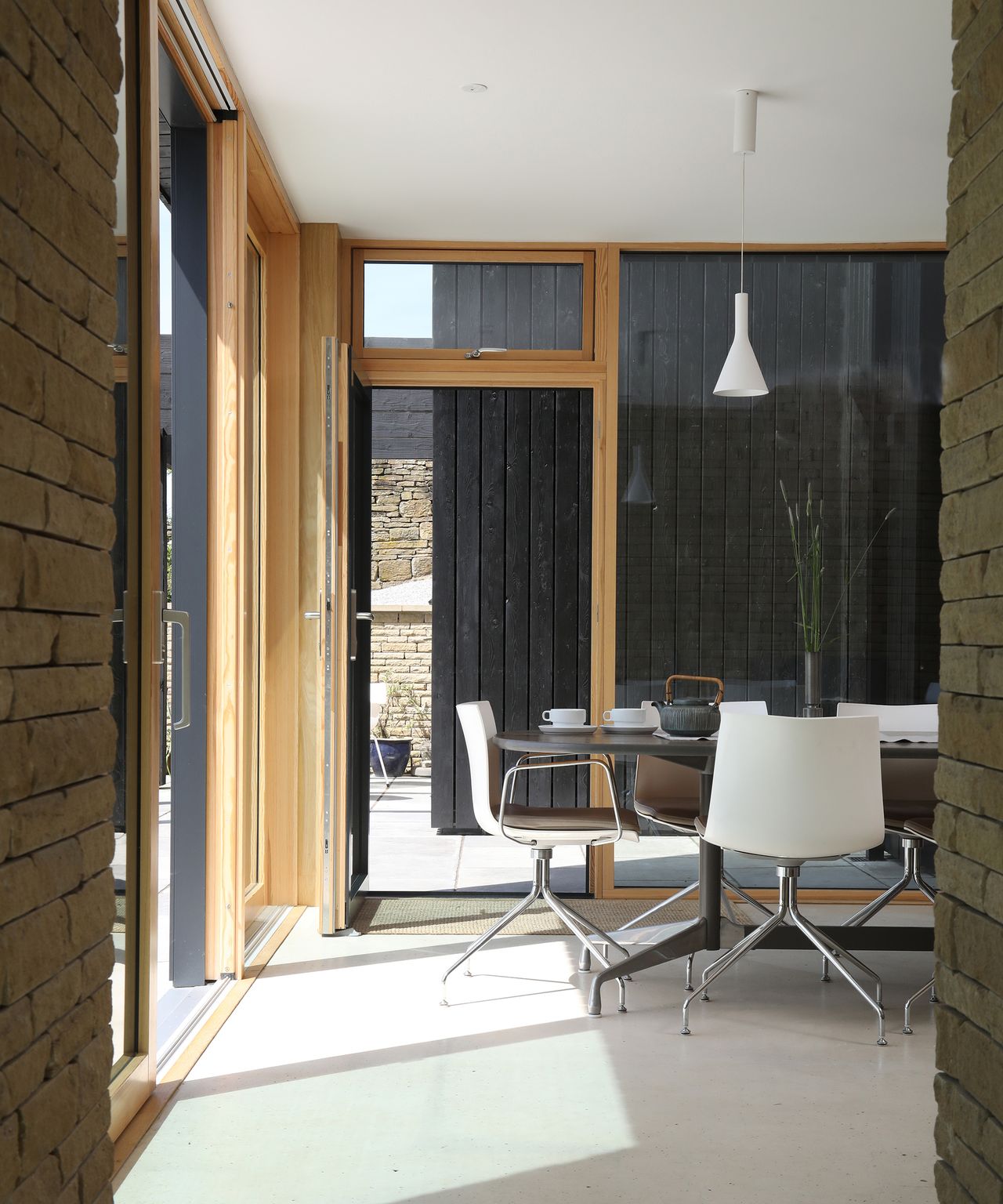 A dining table in a self build home with large glazing which looks through to a second building clad in black timber