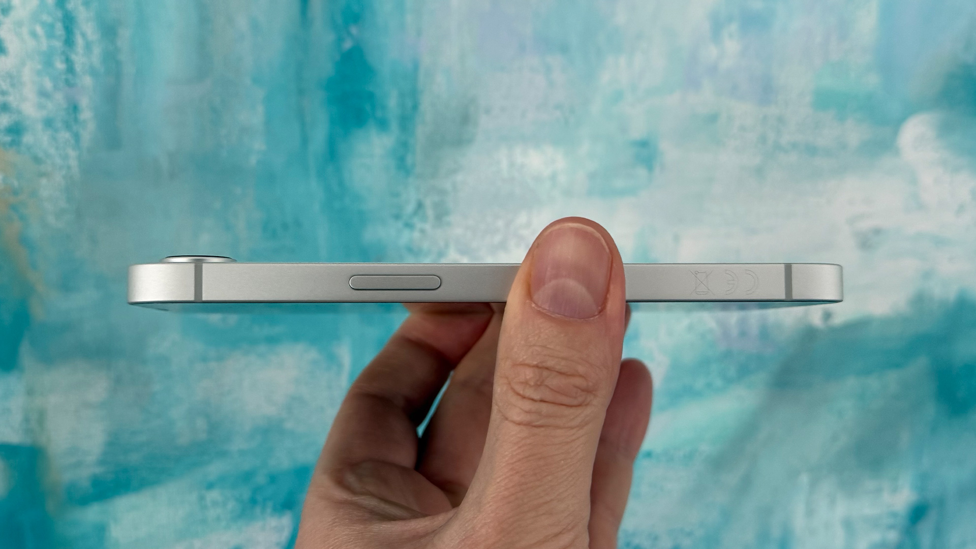 Apple iPhone 16e smartphone held in hand showing side of phone in front of canvas painting