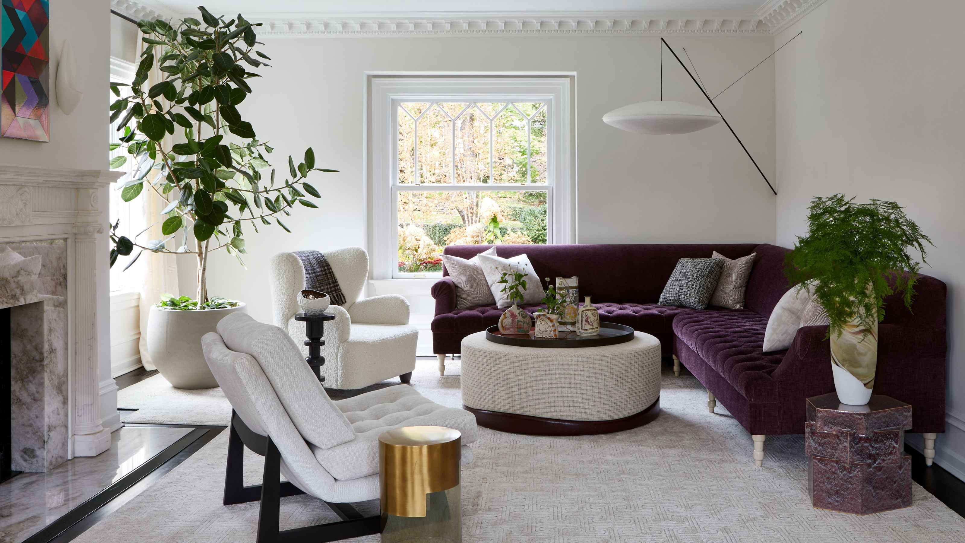 Living room with white walls and a large purple sectional in the right hand corner. A statement wall light hangs above. A white upholstered ottoman sits in the middle and two white armchairs are opposite. A marble fireplace can be seen in the corner