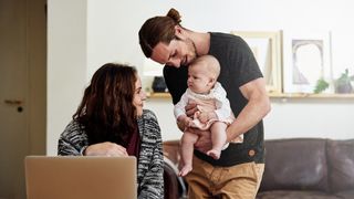 A woman sits in front of the laptop, smiling at her baby as they're being held in her partner's arms