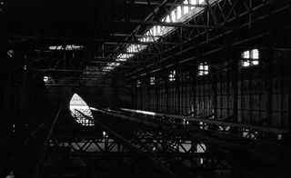 Dark black and white image - looking down the length of interior of a large cladded iron grid-like structure - light shines through an opening at the far end