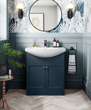 A stylish bathroom featuring a navy blue vanity unit with shaker-style doors and chrome knobs, topped with a sleek white semi-recessed basin. The walls are adorned with a tropical leaf-patterned wallpaper in blue and white, framed by light blue panelling that creates a cohesive and elegant look.