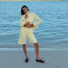 Sandra wearing a yellow jacket and matching Bermuda shorts with black loafers on a white sand beach. 