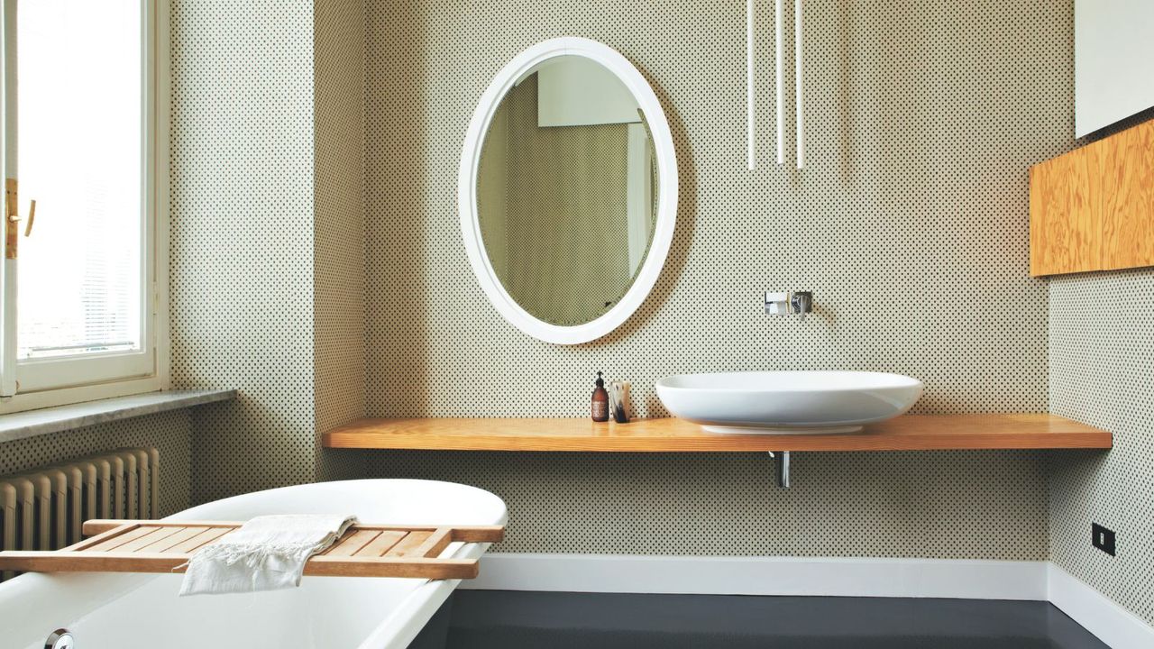 A bright bathroom with a large window, roll top bathtub, wooden counter, spotted wallpaper and large oval mirror