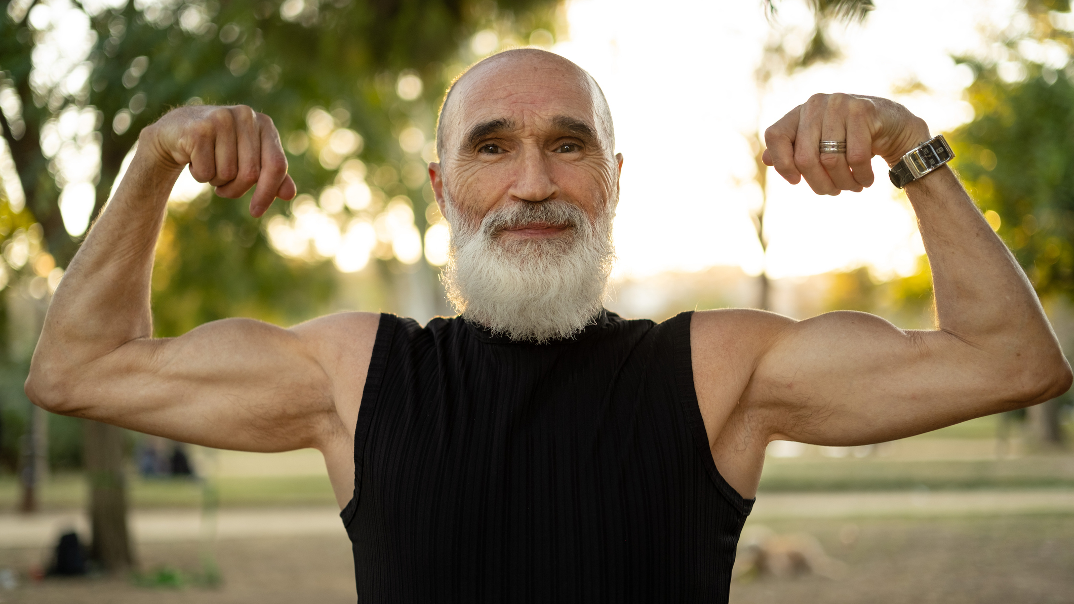 Senior man flexing his biceps. He is outside in a park