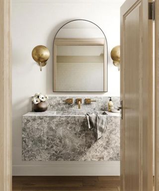 Gold bathroom sconce lights on either side of an arched bathroom mirror. Below, is a floating stone statement sink with gold hardware