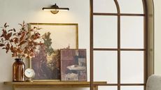 Hearth & Hand™ with Magnolia battery cordless picture light hung in an entryway over a wooden table with stacked artwork and a vase with autumnal foliage