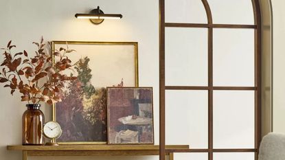 Hearth & Hand™ with Magnolia battery cordless picture light hung in an entryway over a wooden table with stacked artwork and a vase with autumnal foliage