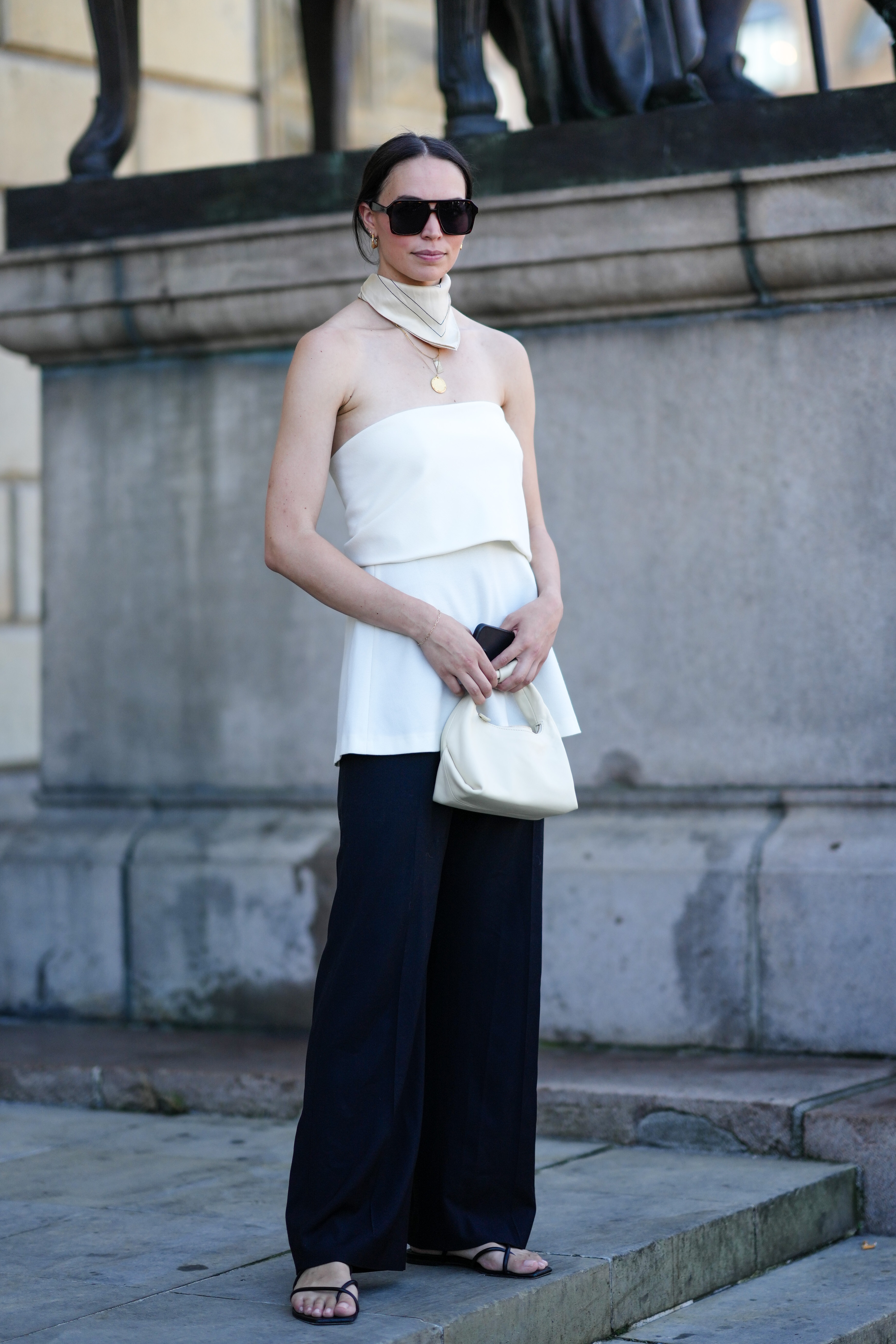 A guest wears black square sunglasses, gold earrings, a beige with black embroidered pattern silk scarf, gold chain pendant necklaces, a white latte shoulder-off / silk top, black wide legs pants, black leather flip flop, a white latte matte leather handbag , outside Helmstedt during Copenhagen Fashion Week Spring/Summer 2023, on August 11, 2022 in Copenhagen, Denmark.