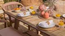 Close-up image of a wooden dining table set with rattan placemats and pink serveware with pink dining chairs around it.