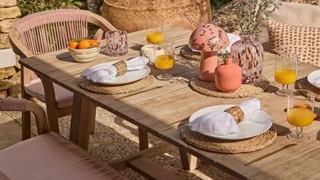 Close-up image of a wooden dining table set with rattan placemats and pink serveware with pink dining chairs around it.