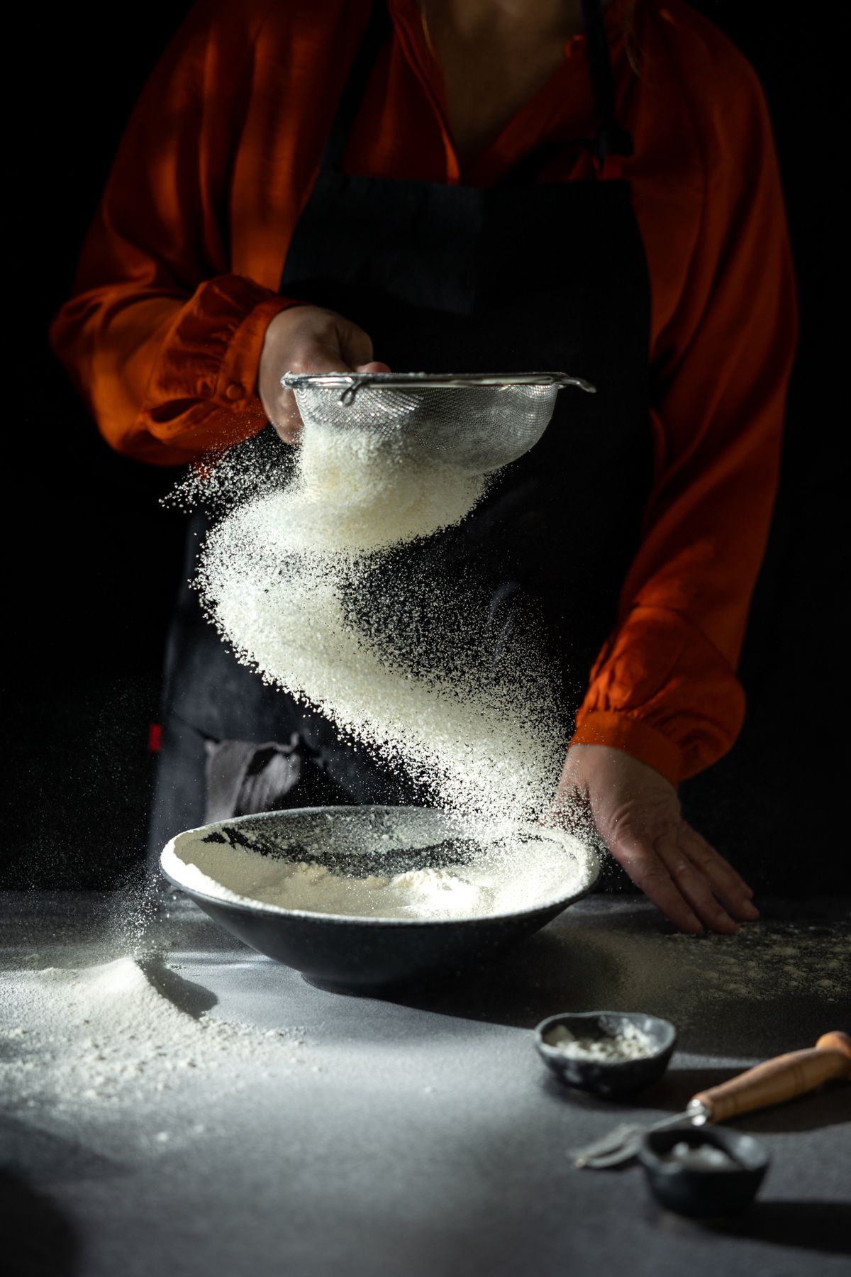 A woman in an orange blouse sifts flour into a black bowl, creating a swirling pattern of flour in the air against a dark background