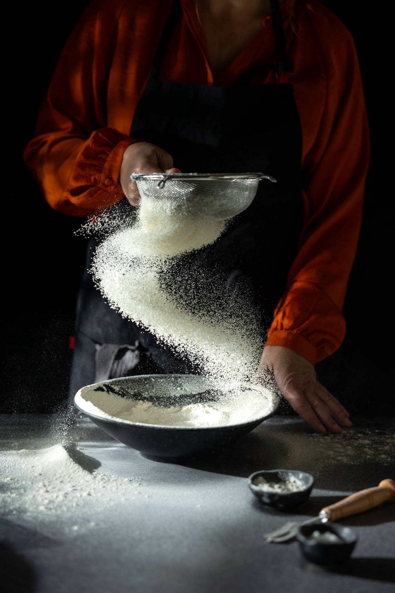 A woman in an orange blouse sifts flour into a black bowl, creating a swirling pattern of flour in the air against a dark background