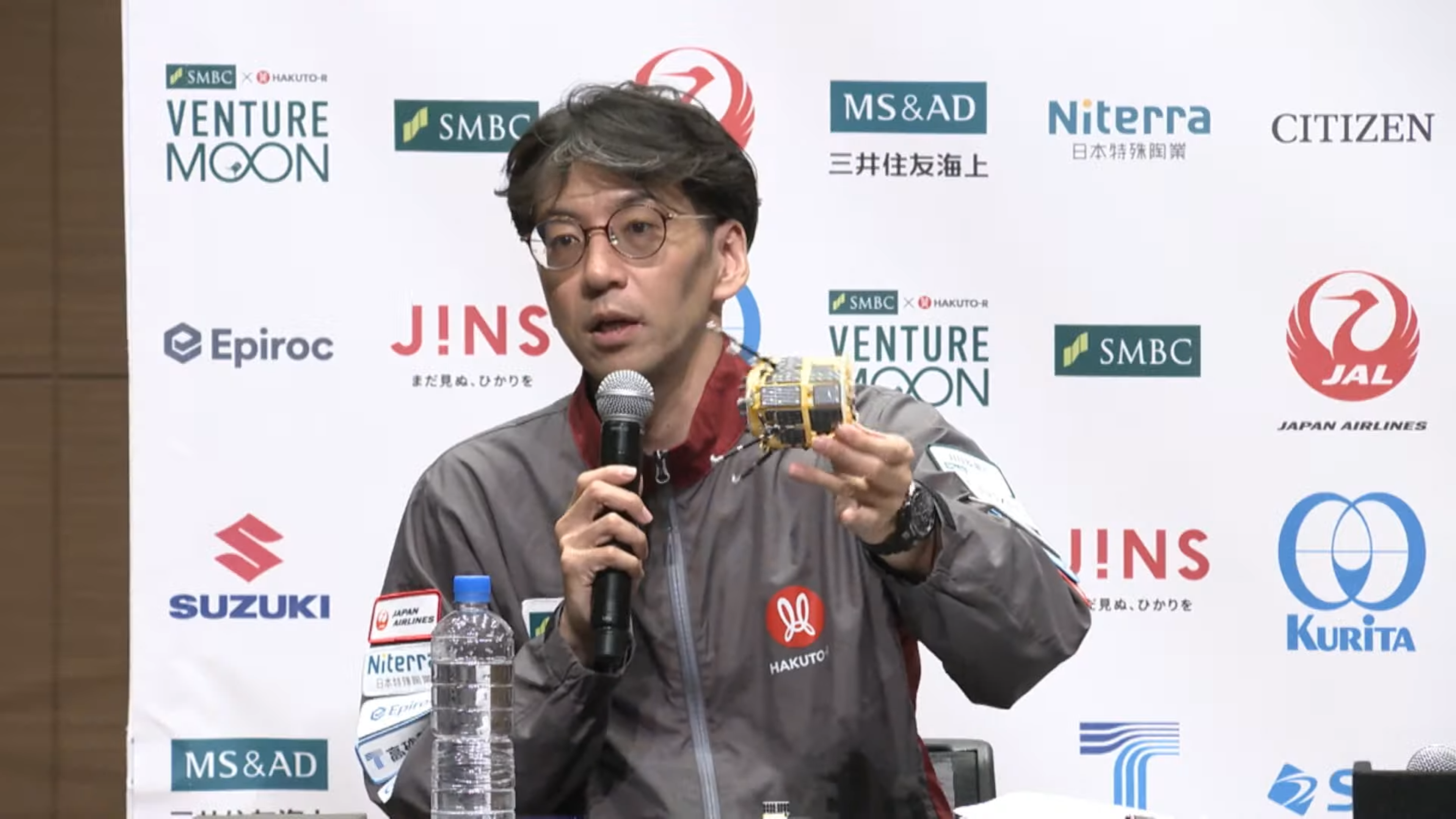 An ispace engineer holds a model of a Resilience moon lander while speaking into a microphone.