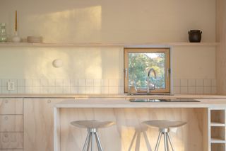 A close up of the kitchen space, with simple white tiling, metal high stools, and timber kitchen