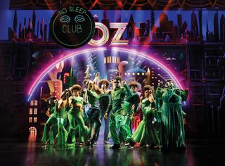 a group of dancers dressed in green costumes pose in front of a city skyline, with a circular neon pink and purple decoration and signs reading no sleep club and oz