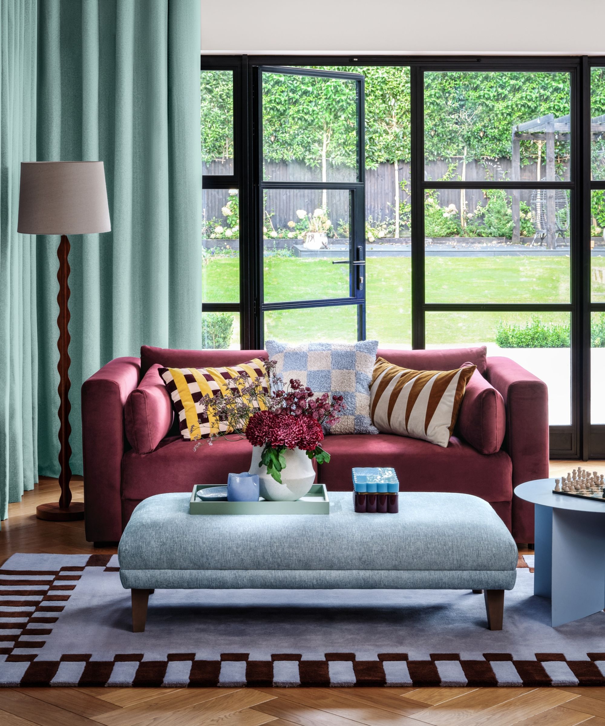 A burgundy colored sofa behind a light blue upholstered ottoman in a bright living room. A large glass door behind overlooking a lawn.
