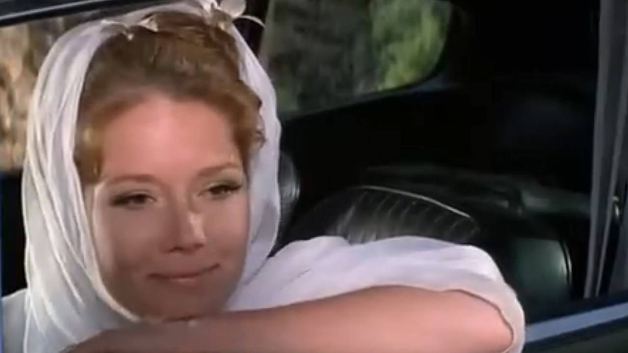 Diana Rigg smiling while sitting in a car.